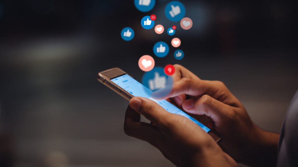 Person typing on phone screen while thumbs up and heart icons float out of it.